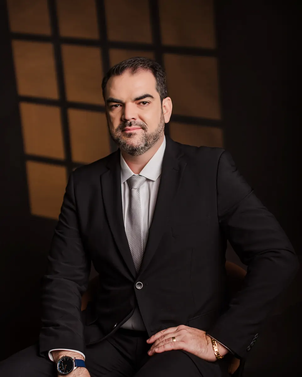 Denisson Rabelo Rebonato sentado, de terno escuro, em frente a uma janela com luz de fim de tarde